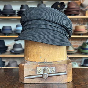 Straw Casquette in black with a textured woven design, displayed on a wooden hat block in a hat shop featuring various styles in the background.