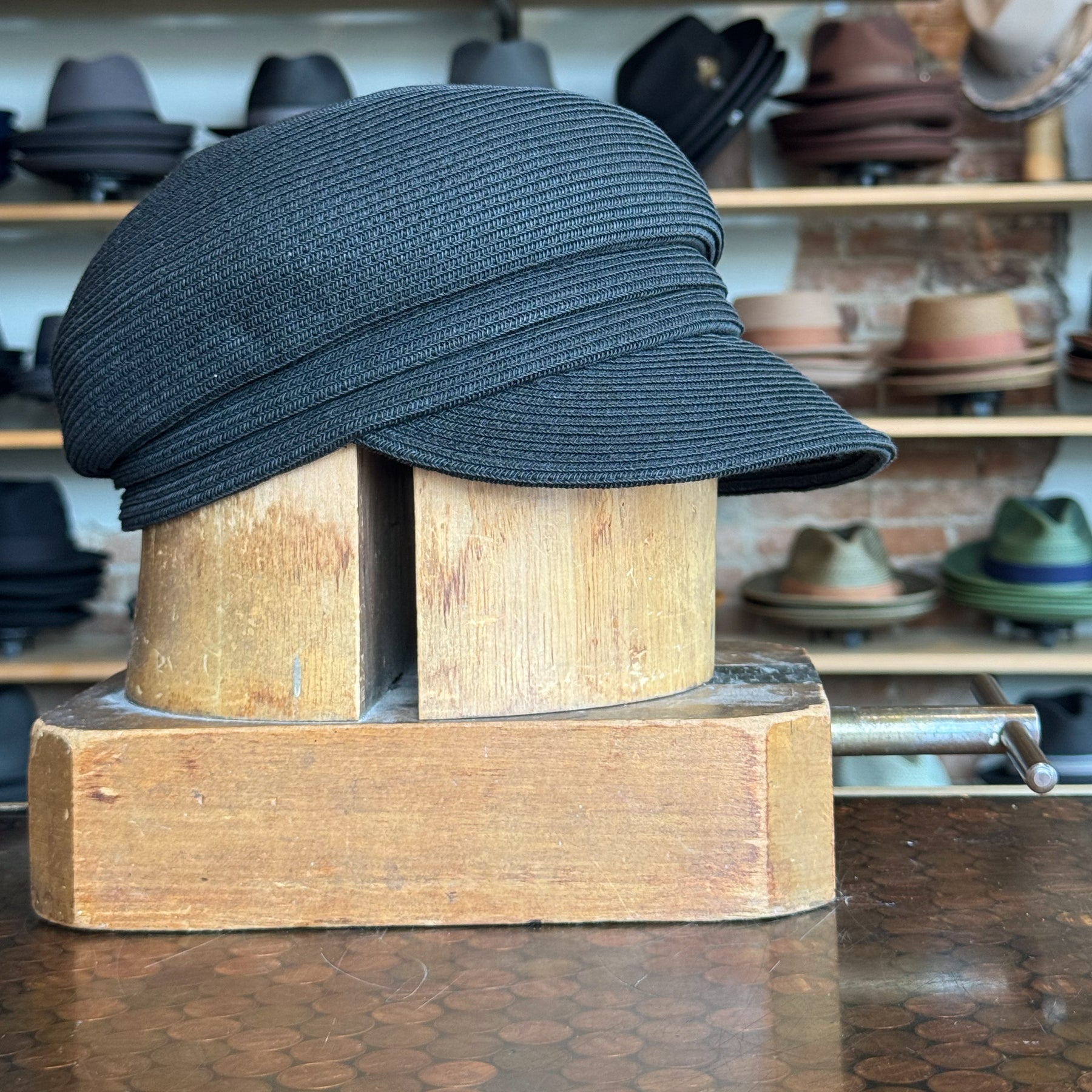 Black Straw Casquette displayed on a wooden hat block in a store setting with other hats blurred in the background, showcasing its structured brim and woven texture.