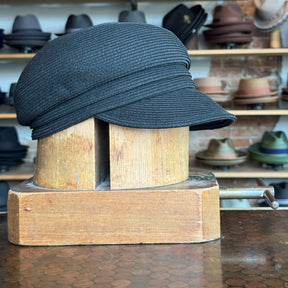 Black Straw Casquette displayed on a wooden hat block in a store setting with other hats blurred in the background, showcasing its structured brim and woven texture.