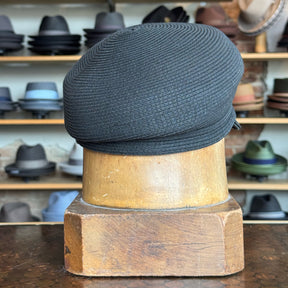 Black woven Straw Casquette displayed on a wooden hat stand in a store with various other hats in the background.
