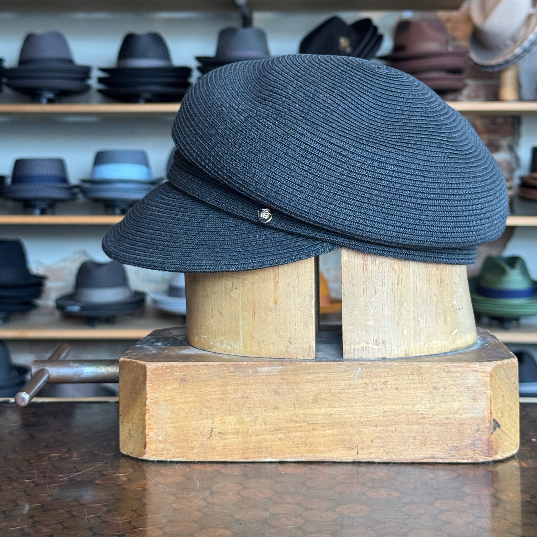 Black Straw Casquette hat with a curved brim and small decorative button, displayed on a wooden hat block in a shop with various hats in the background.