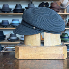 Black Straw Casquette hat with a curved brim and small decorative button, displayed on a wooden hat block in a shop with various hats in the background.