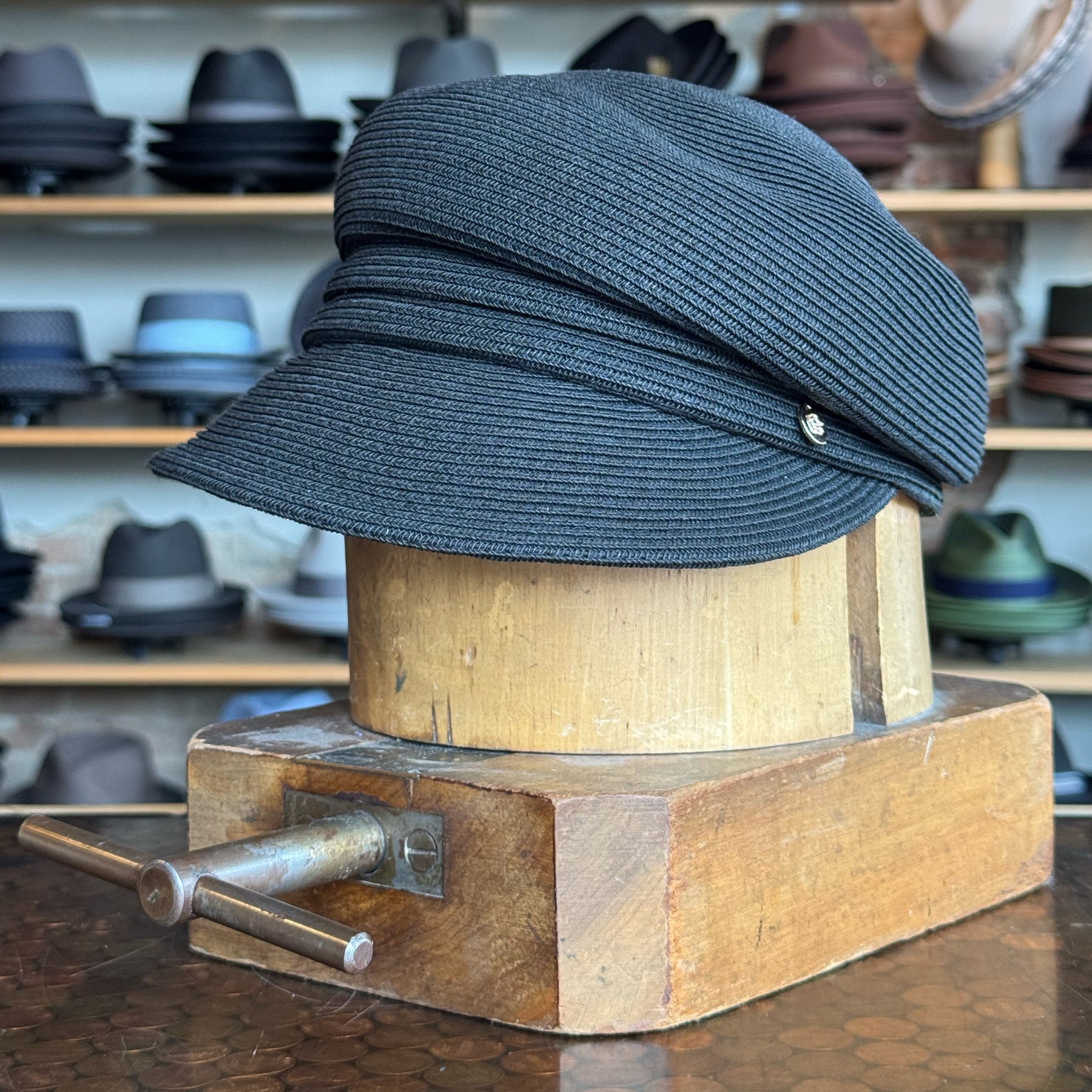Straw Casquette in black displayed on a wooden hat block, showcasing its textured woven material and classic newsboy cap style with a curved brim.