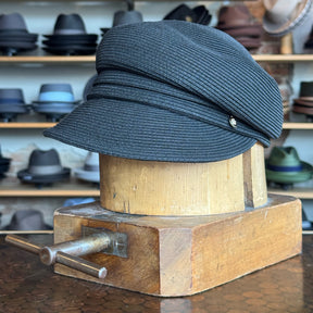 Straw Casquette in black displayed on a wooden hat block, showcasing its textured woven material and classic newsboy cap style with a curved brim.