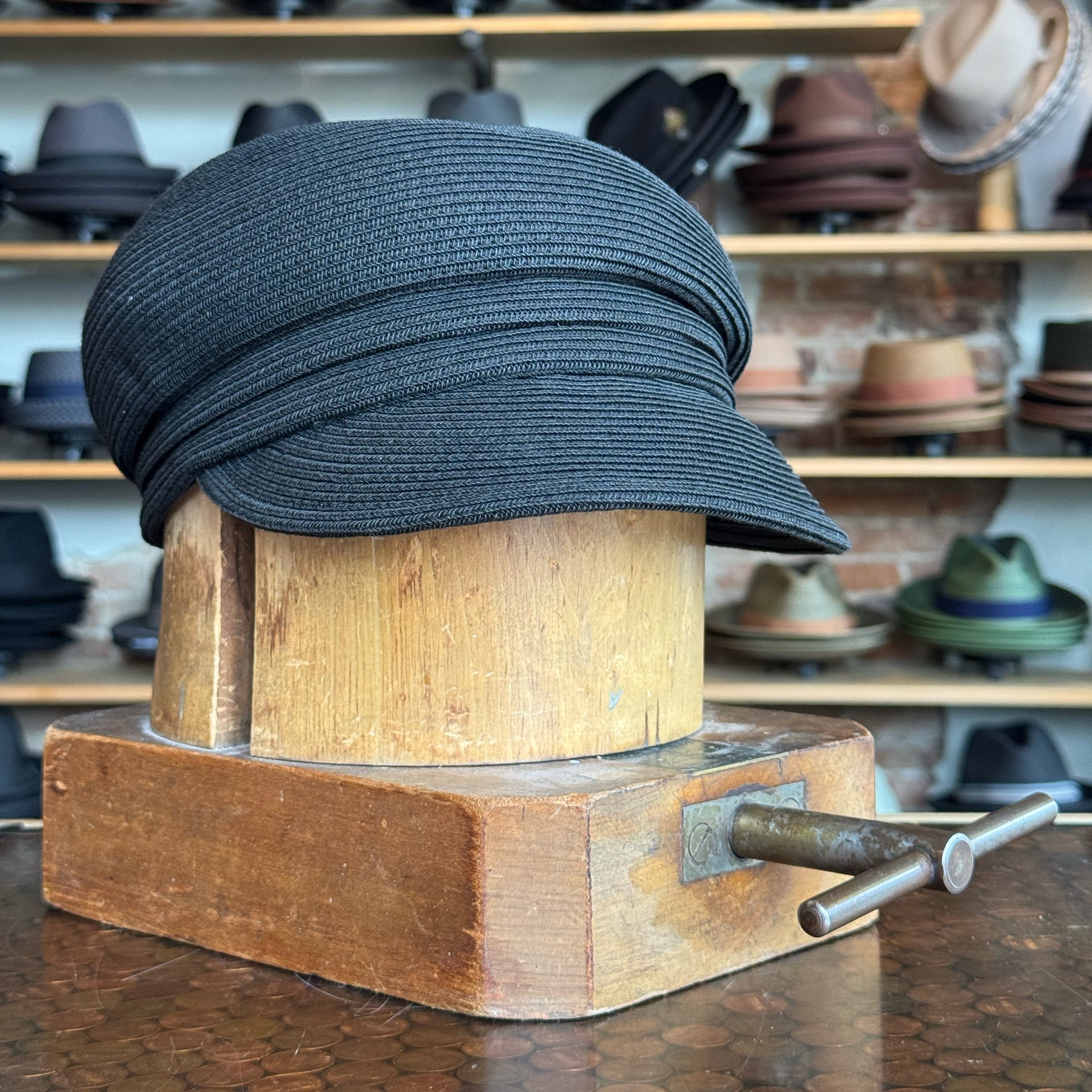 Black Straw Casquette cap displayed on a vintage wooden hat block in a shop with various hats in the background, showcasing a stylish newsboy design.