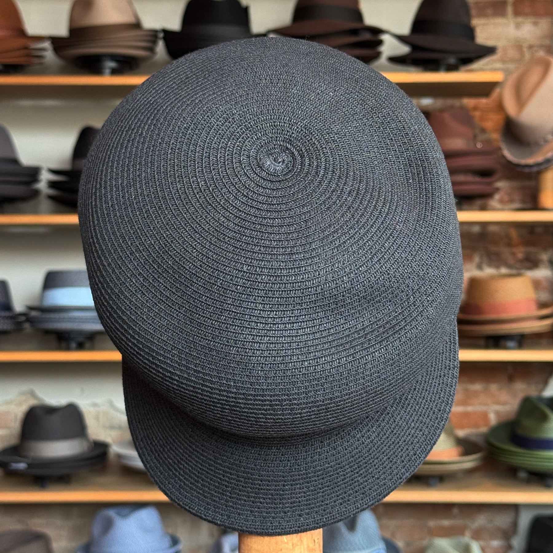Black woven Straw Casquette with a flat brim displayed in a hat shop with various hats in the background