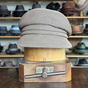 Straw Casquette in a light milk chocolate color displayed on a wooden hat block, surrounded by various hats in the background, showcasing a stylish woven newsboy cap design.