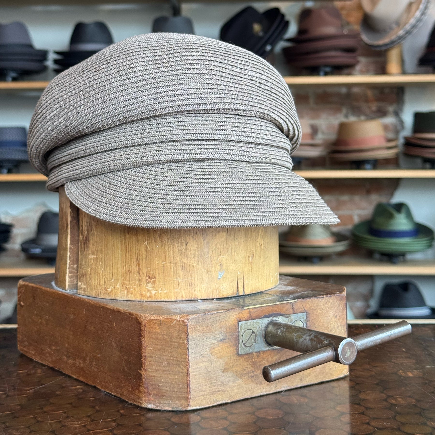 Straw Casquette in a soft brown tone displayed on a vintage wooden hat block, surrounded by various hats on shelves in the background.