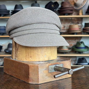 Straw Casquette in a soft brown tone displayed on a vintage wooden hat block, surrounded by various hats on shelves in the background.