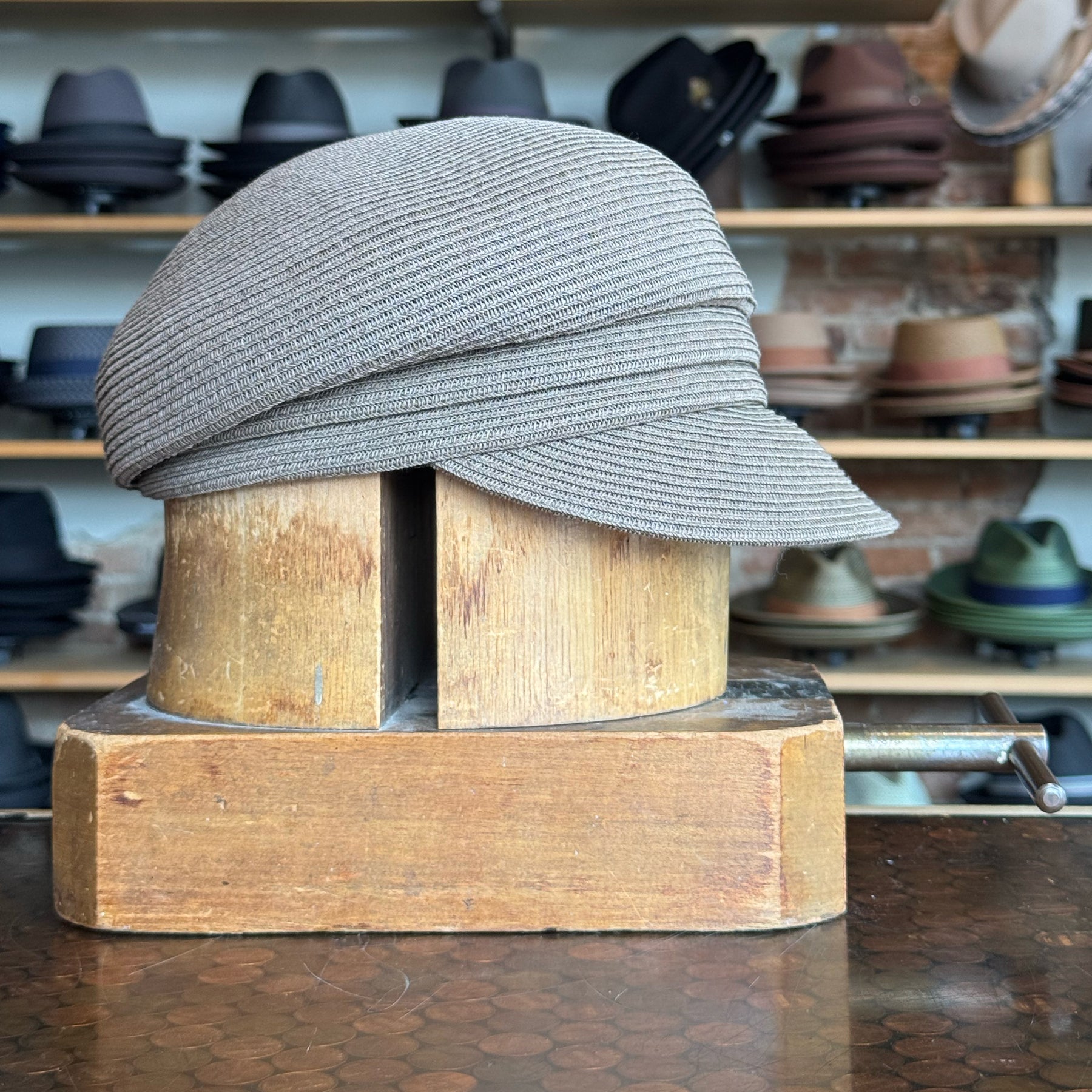 Straw Casquette in a soft beige tone displayed on a wooden hat block with shelves of various hats in the background, showcasing a stylish woven accessory for casual wear.