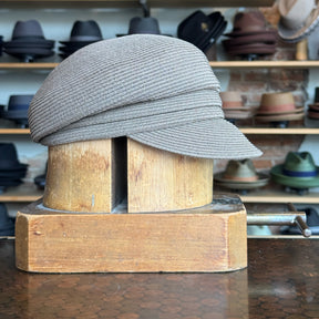 Straw Casquette in a soft beige tone displayed on a wooden hat block with shelves of various hats in the background, showcasing a stylish woven accessory for casual wear.