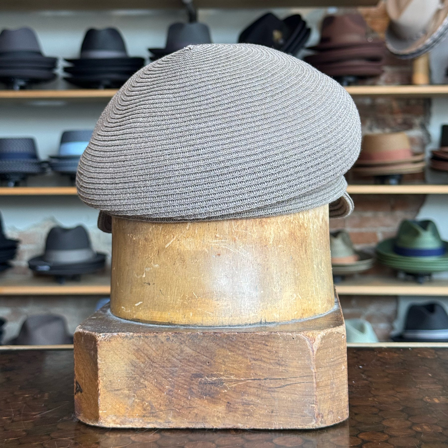 Straw Casquette hat in a soft milk chocolate color displayed on a vintage wooden hat block with a background shelving of various hats including fedoras and newsboy caps.