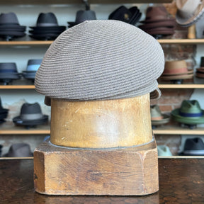 Straw Casquette hat in a soft milk chocolate color displayed on a vintage wooden hat block with a background shelving of various hats including fedoras and newsboy caps.