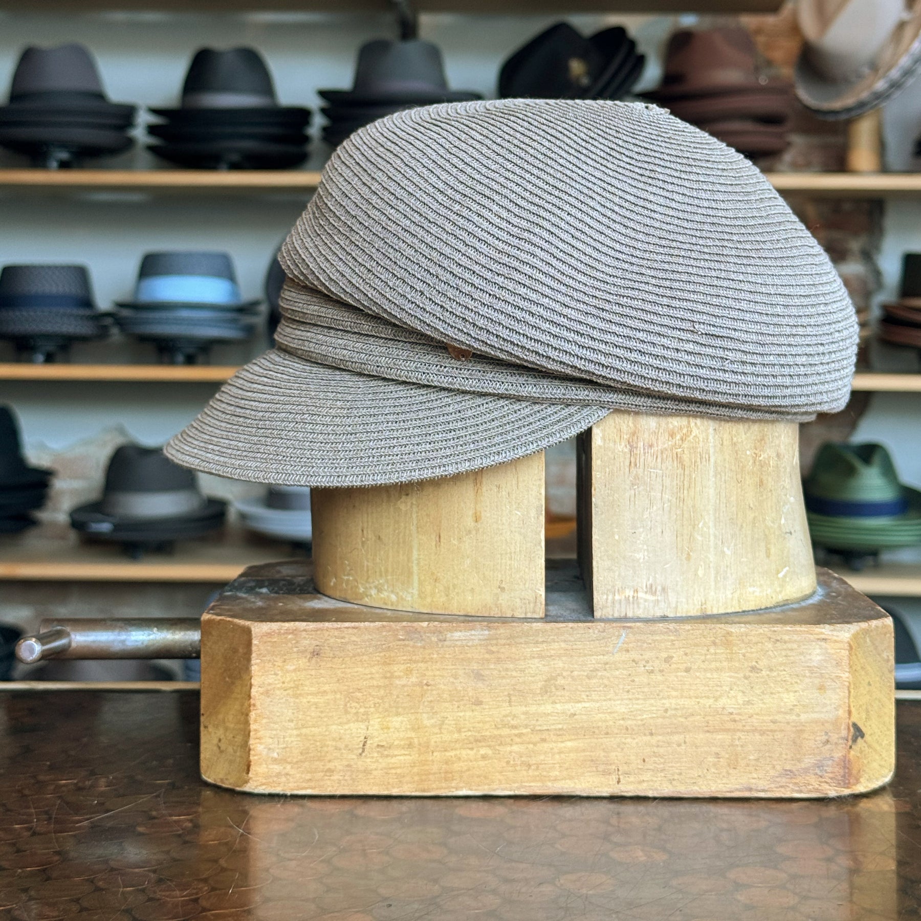 Straw Casquette displayed on a wooden hat block in front of shelves filled with various hats, showcasing its light brown woven texture and classic flat cap style.