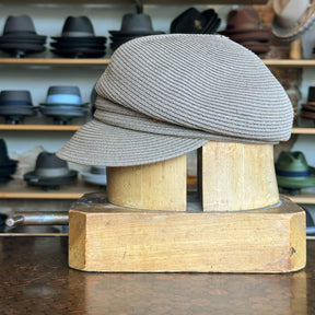 Straw Casquette displayed on a wooden hat block in front of shelves filled with various hats, showcasing its light brown woven texture and classic flat cap style.