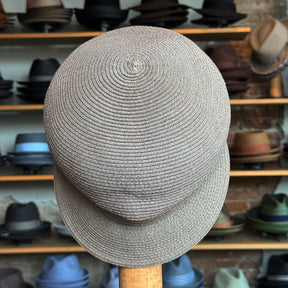 Straw Casquette in a light natural straw color, woven with a classic rounded crown and structured brim, displayed in a hat shop with various styles in the background.