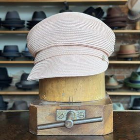 Straw Casquette in soft pink with a textured weave, featuring a structured brim and classic newsboy cap style, displayed on a wooden hat stand in a hat shop.