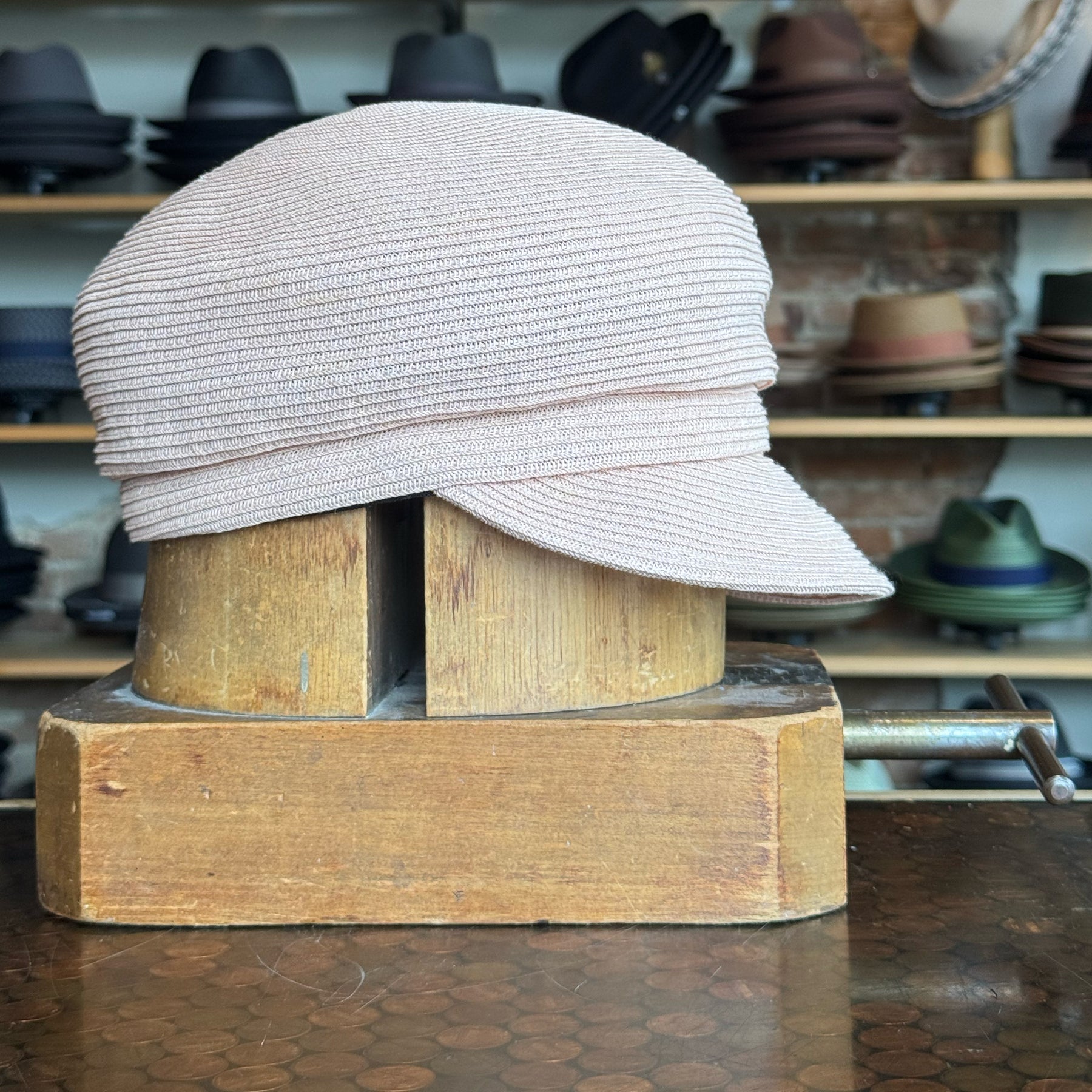 Straw Casquette in soft pink displayed on a wooden hat block, showcasing a textured woven design and short brim, surrounded by various hats on shelves in the background.