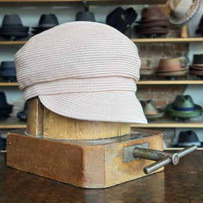 Light pink Straw Casquette with a textured woven design displayed on a wooden hat block in a hat shop featuring various hats in the background.
