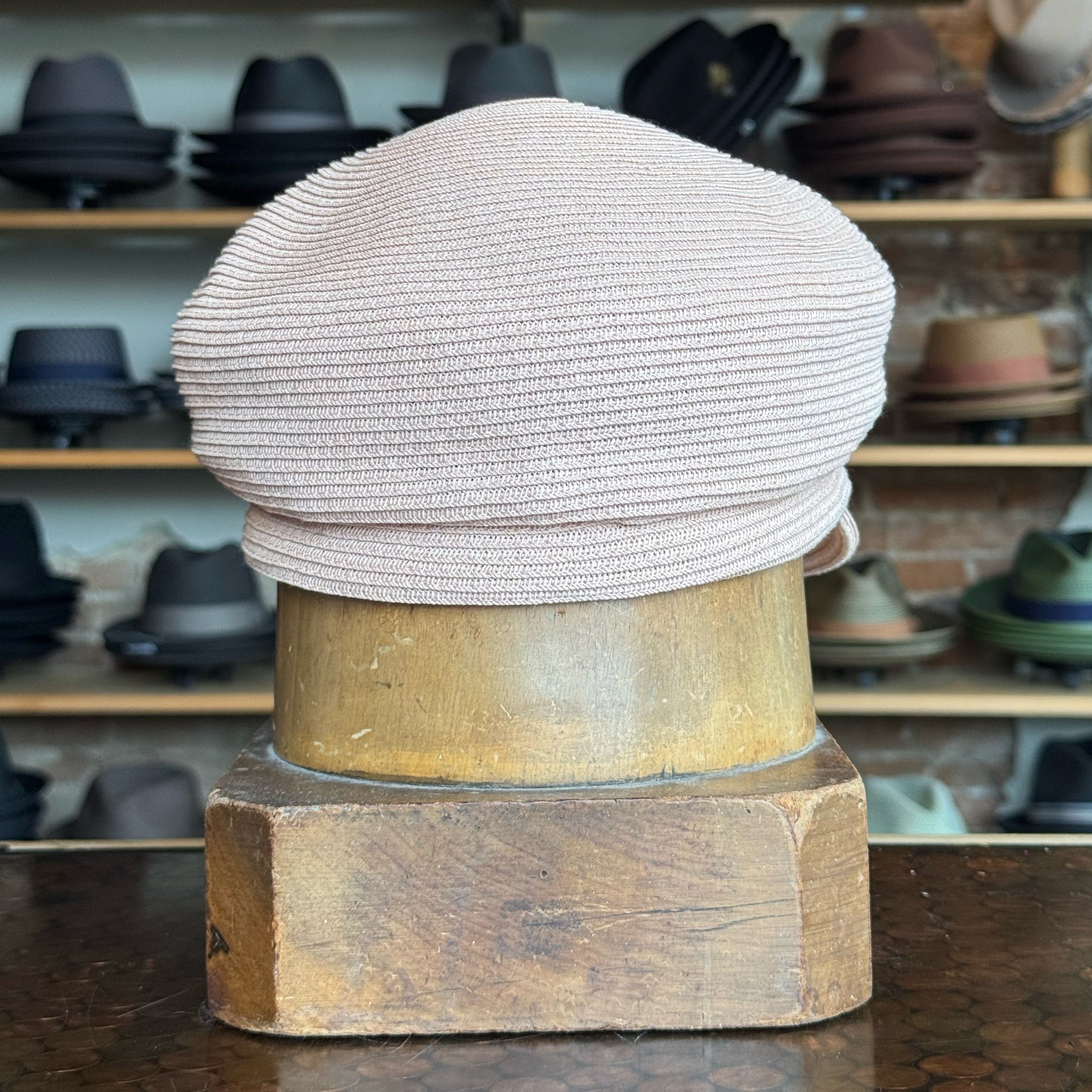 Light pink woven Straw Casquette displayed on a wooden hat block with various hats in the background, showcasing a classic newsboy cap style.