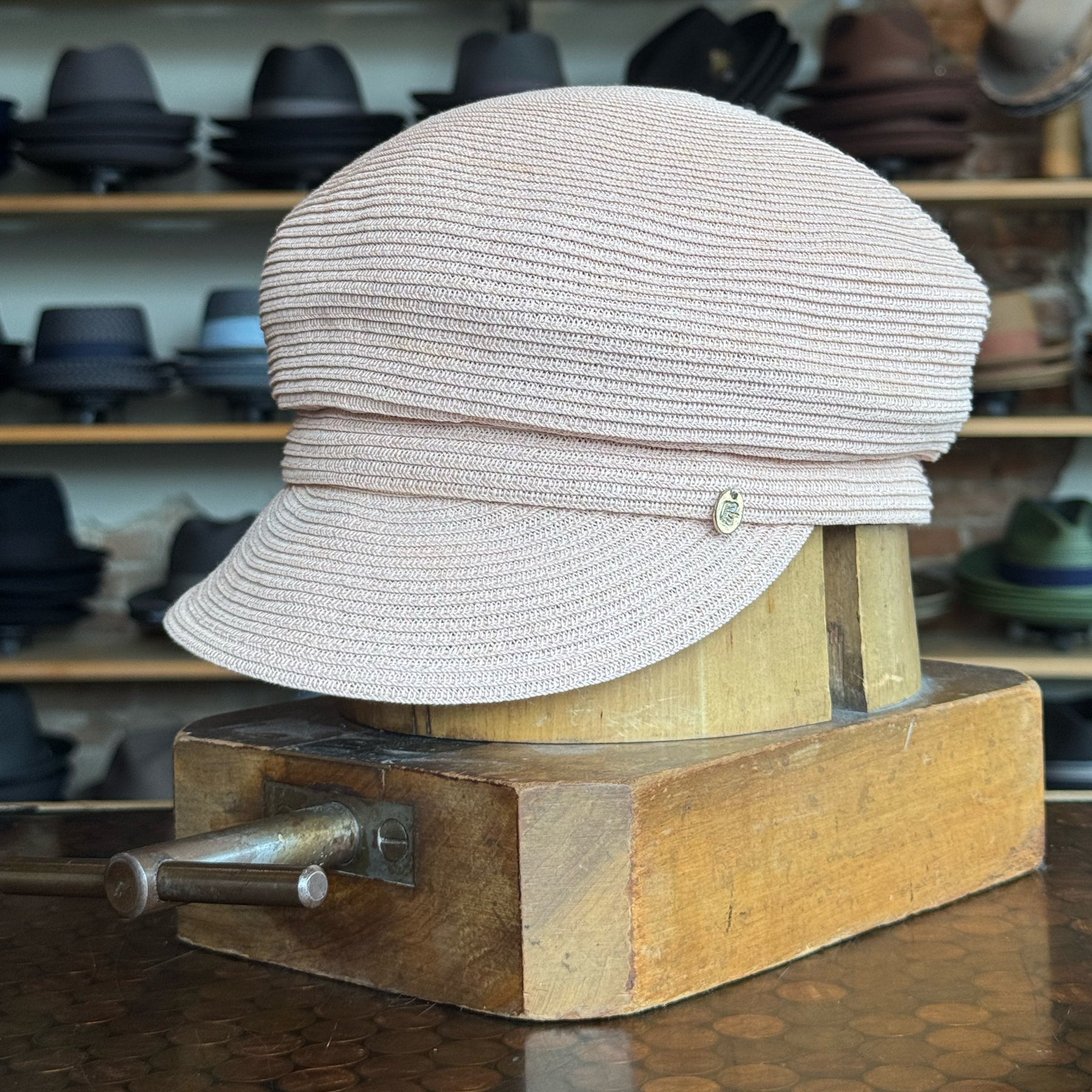 Light pink straw casquette with a curved brim and textured woven design, displayed on a wooden stand in a shop with various hats in the background.