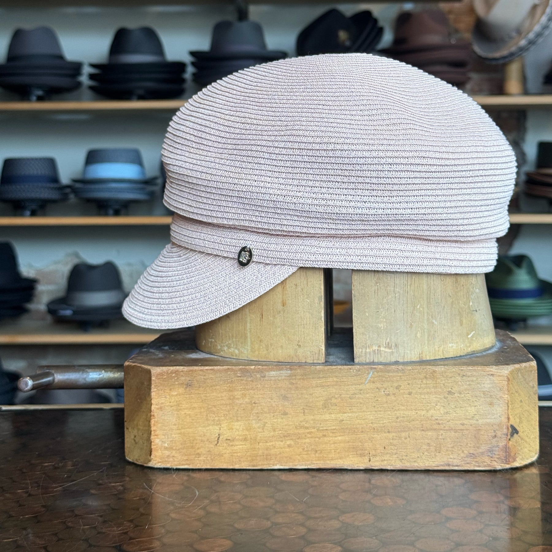 Light pink woven Straw Casquette with a small side emblem displayed on a wooden hat block against a background of various hats on shelves.