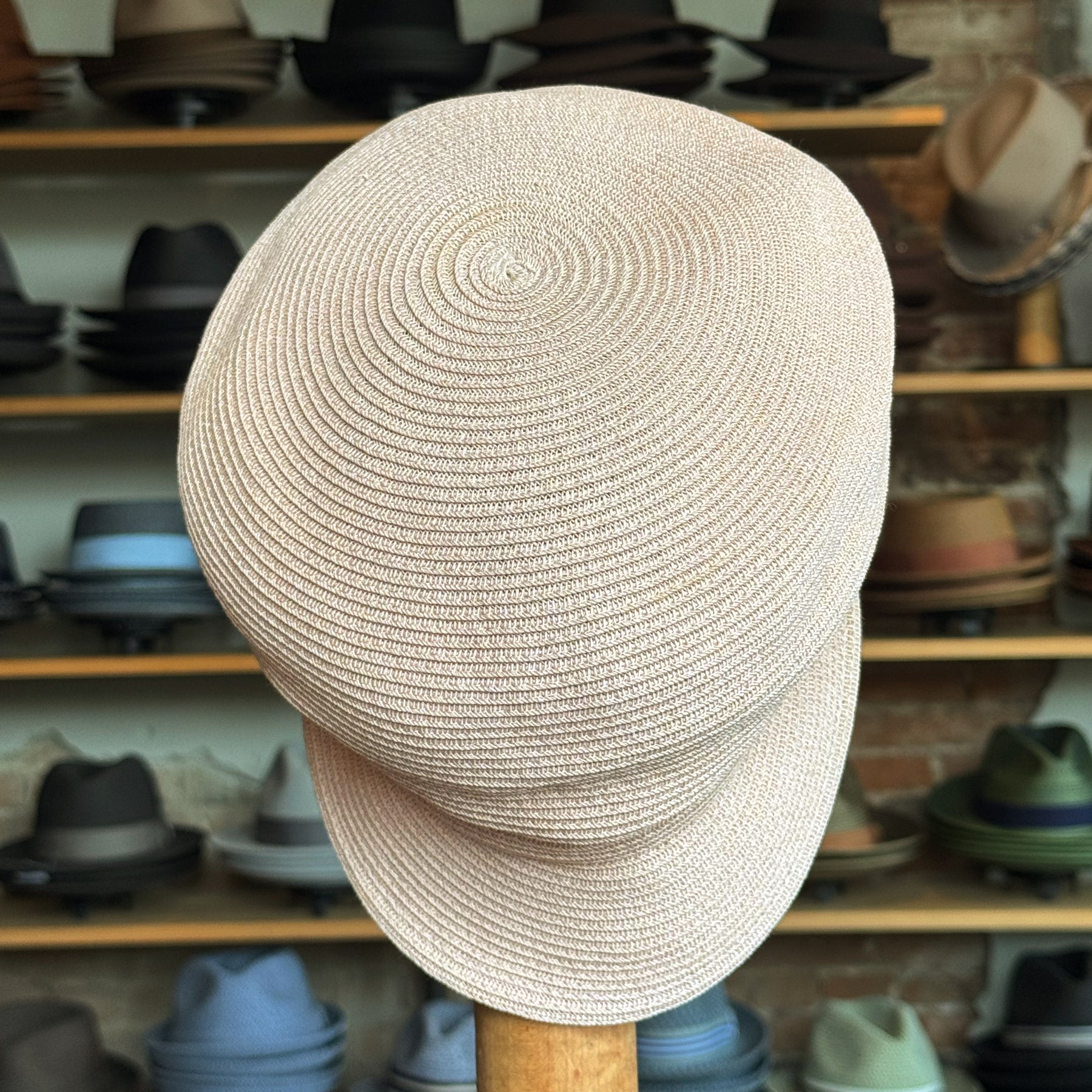 Straw Casquette in a light natural color with a woven texture, displayed on a stand in a hat shop featuring various caps and hats in the background.