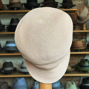 Straw Casquette in a light natural color with a woven texture, displayed on a stand in a hat shop featuring various caps and hats in the background.