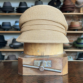 Straw Casquette in natural beige color displayed on a wooden hat block, styled with a curved brim and soft crown, surrounded by various hats in the background.