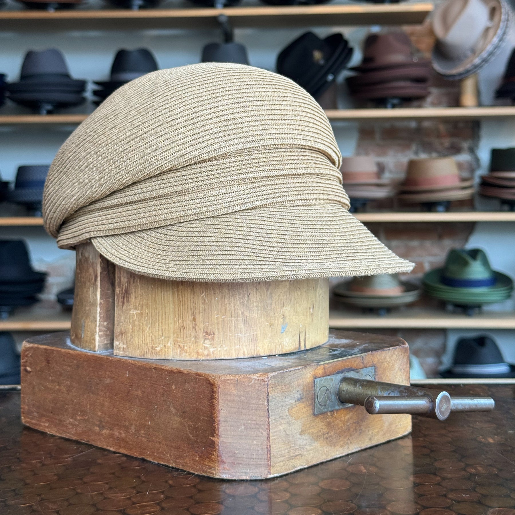 Light tan Straw Casquette hat with a short brim displayed on a wooden hat block, surrounded by various hats on shelves in a store setting.