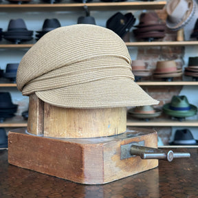 Light tan Straw Casquette hat with a short brim displayed on a wooden hat block, surrounded by various hats on shelves in a store setting.