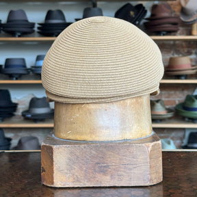 Straw Casquette in a light tan color displayed on a wooden hat block, with shelves of various styles of hats blurred in the background.