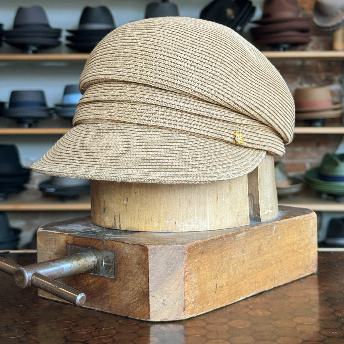 Straw Casquette displayed on a wooden hat block, featuring a woven natural straw texture with a soft, structured design and a small metallic accent on the side.