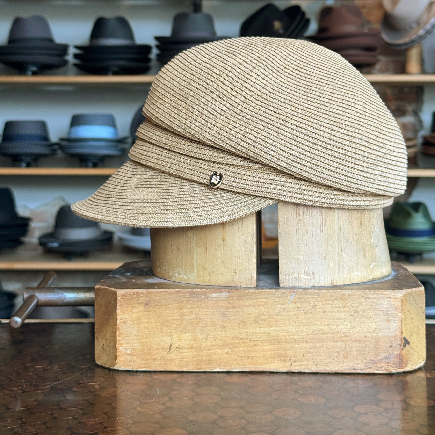 Beige Straw Casquette with a wrap-around band detail and small decorative pin, displayed on a wooden hat stand in a shop with various hats in the background.