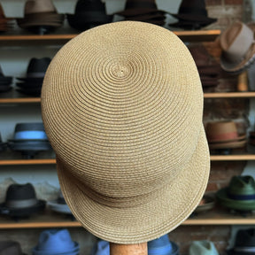 Straw Casquette made from woven natural straw fibers with a rounded crown and short, structured brim, displayed against a backdrop of various hats on wooden shelves.