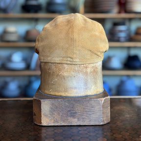 Back view of a cognac-colored Summer Suede 6-Panel Cap displayed on a wooden hat block with shelves of various hats in the background.