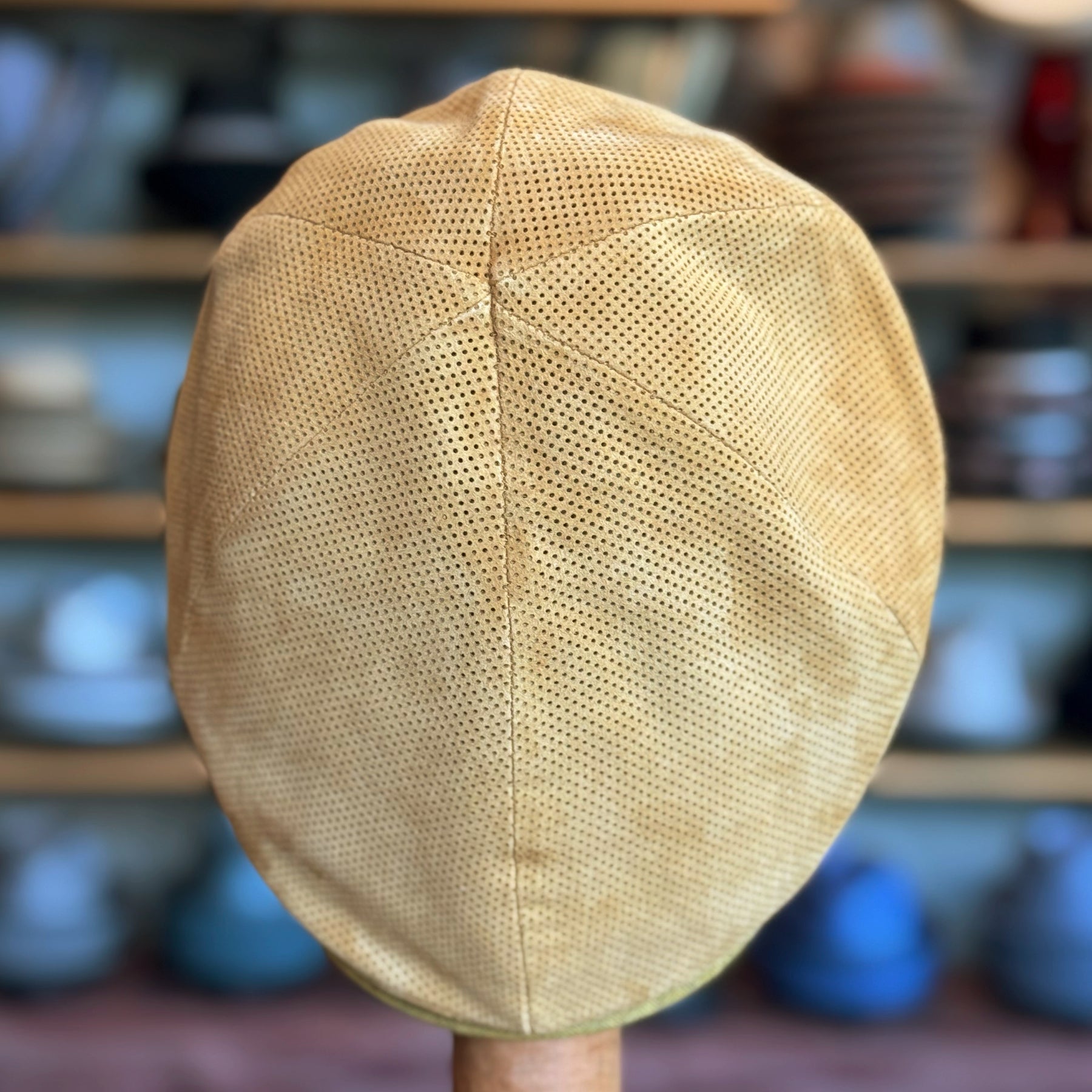 Back view of a tan perforated suede Summer Suede 6-Panel Cap displayed on a wooden stand with a blurred background of shelves holding various hats and caps.