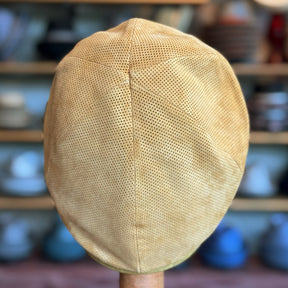 Back view of a tan perforated suede Summer Suede 6-Panel Cap displayed on a wooden stand with a blurred background of shelves holding various hats and caps.