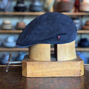 Summer Suede 6-Panel Cap in navy blue suede material displayed on a wooden hat block, showcasing its classic flat cap style with textured fabric and subtle detailing.