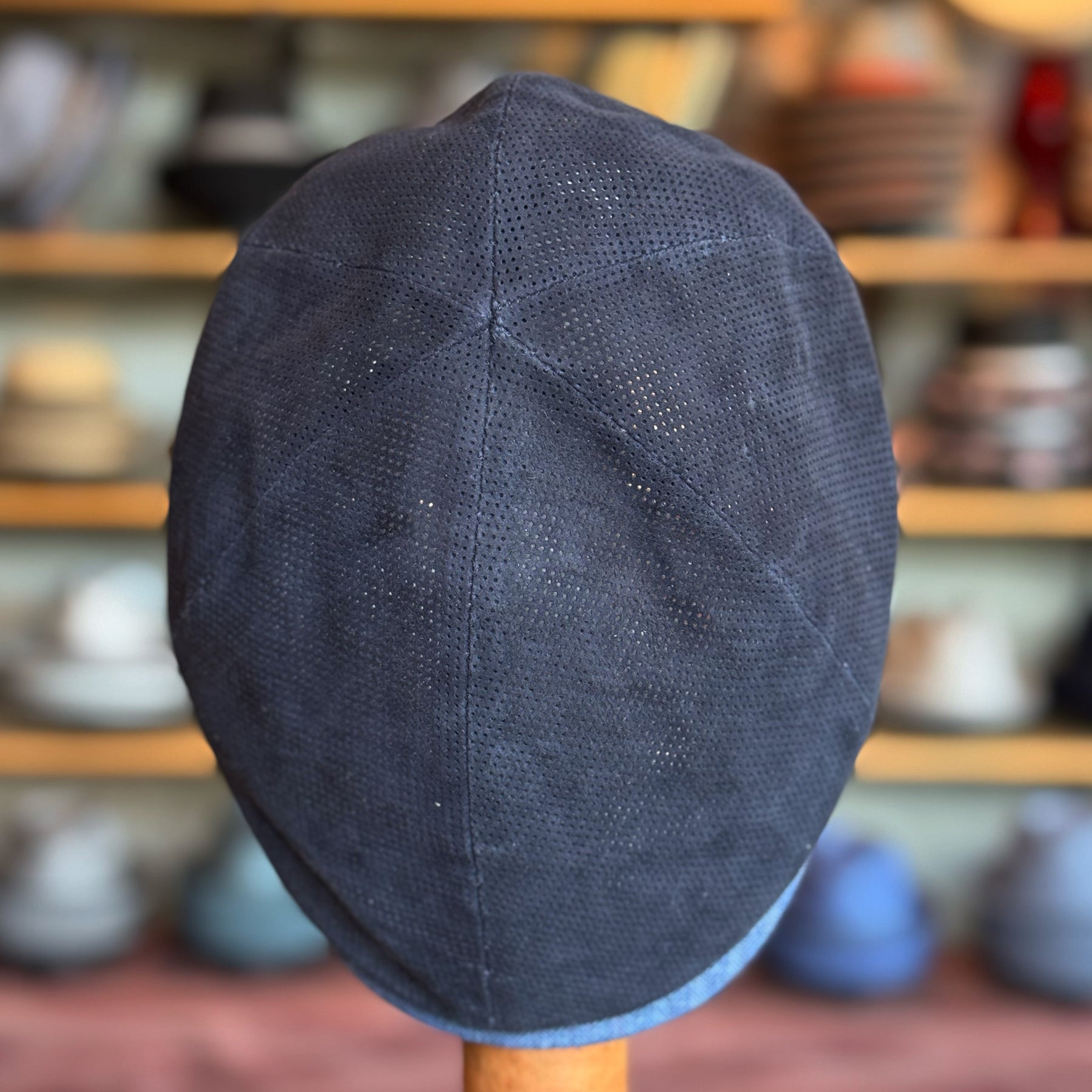 Back view of a navy blue Summer Suede 6-Panel Cap made from perforated suede material, displayed on a stand with blurred shelves of additional hats in the background.