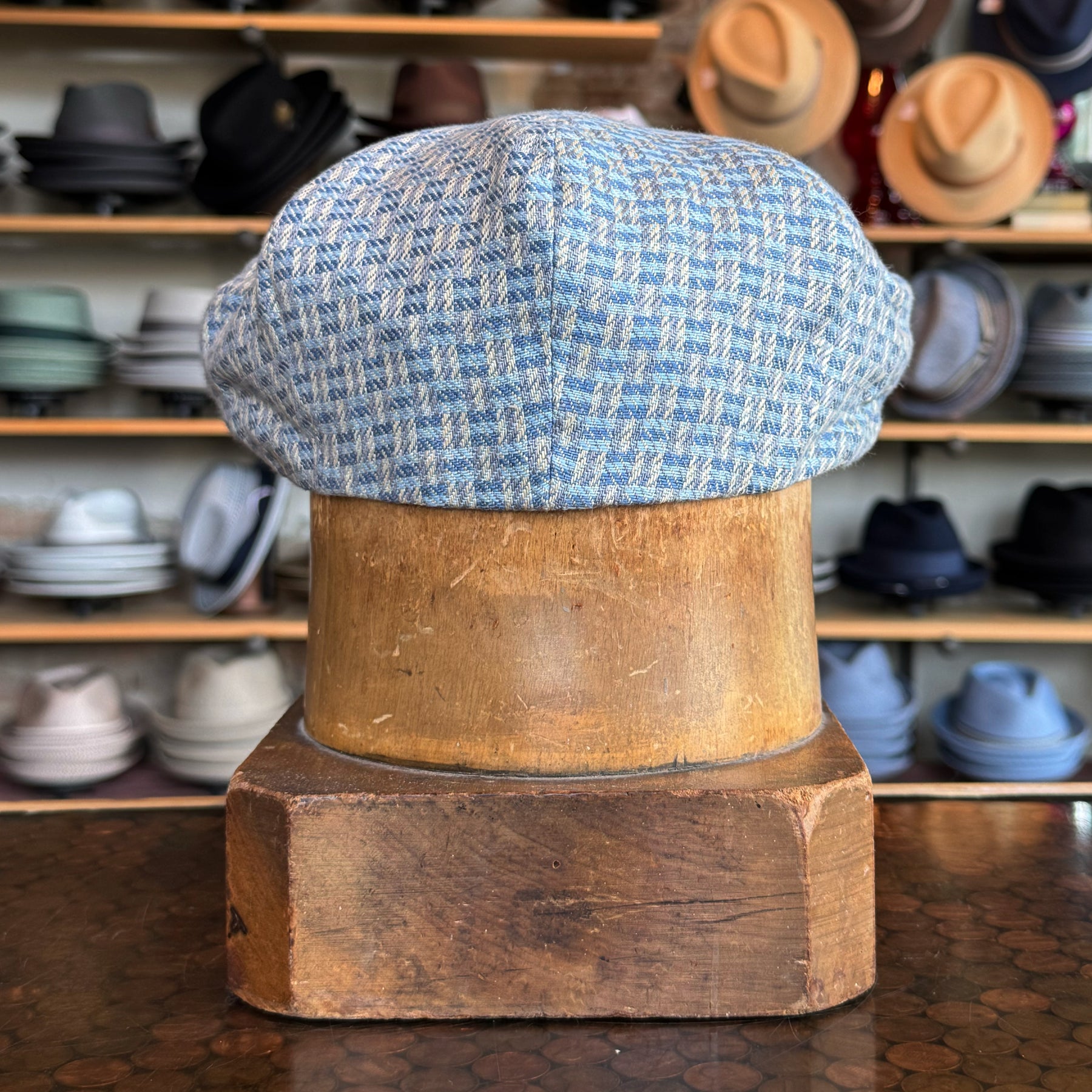 Light blue and beige patterned fabric Tetris Jacquard Newsboy cap displayed on a wooden hat block with shelves of various hats in the background