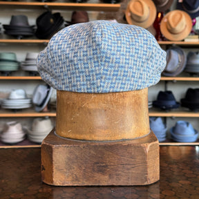 Light blue and beige patterned fabric Tetris Jacquard Newsboy cap displayed on a wooden hat block with shelves of various hats in the background