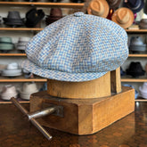 Tetris Jacquard Newsboy cap in a soft blue and cream pattern displayed on a wooden hat block, surrounded by various hats in the background, showcasing a classic newsboy style.
