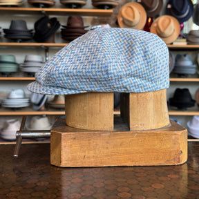 Side view of a light blue and beige patterned Tetris Jacquard Newsboy cap displayed on a wooden hat block against a backdrop of various hats on shelves in a shop.
