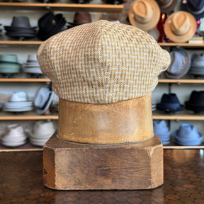 Back view of a beige and gold woven flat cap displayed on a wooden hat stand, showcasing the Tetris Jacquard Newsboy style in a shop setting with various hats in the background.