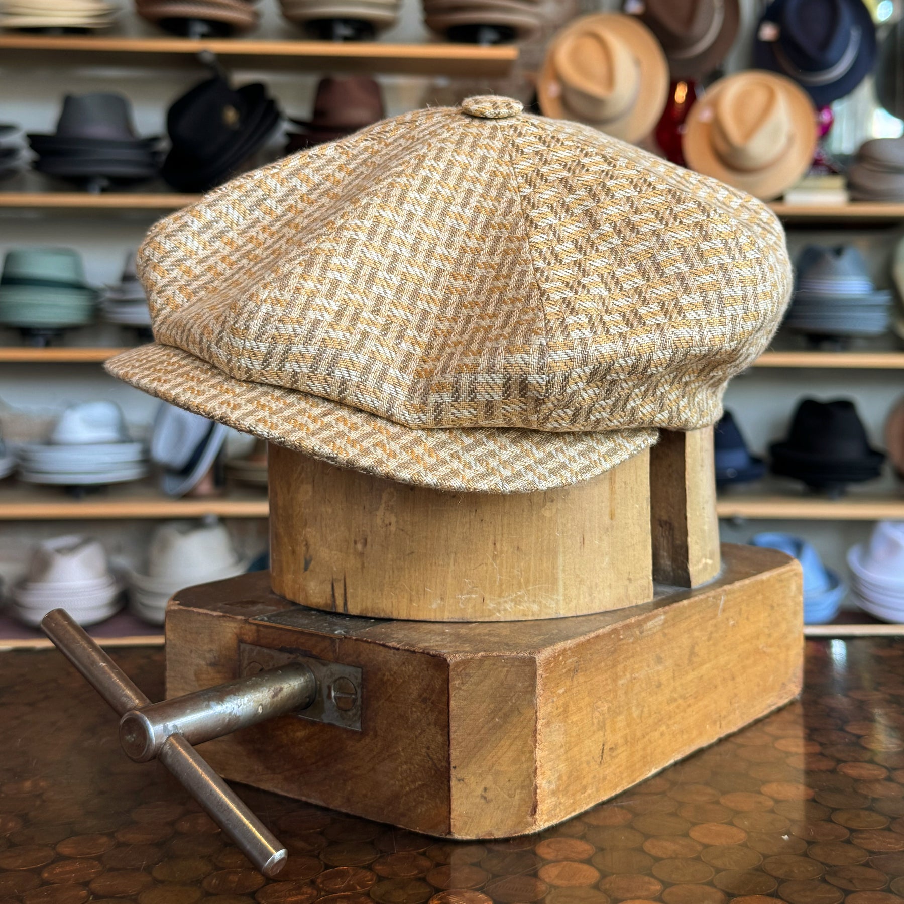 Tetris Jacquard Newsboy cap in a beige and gold woven pattern displayed on a wooden block with various hats blurred in the background in a hat shop.