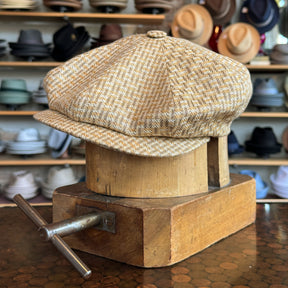 Tetris Jacquard Newsboy cap in a beige and gold woven pattern displayed on a wooden block with various hats blurred in the background in a hat shop.