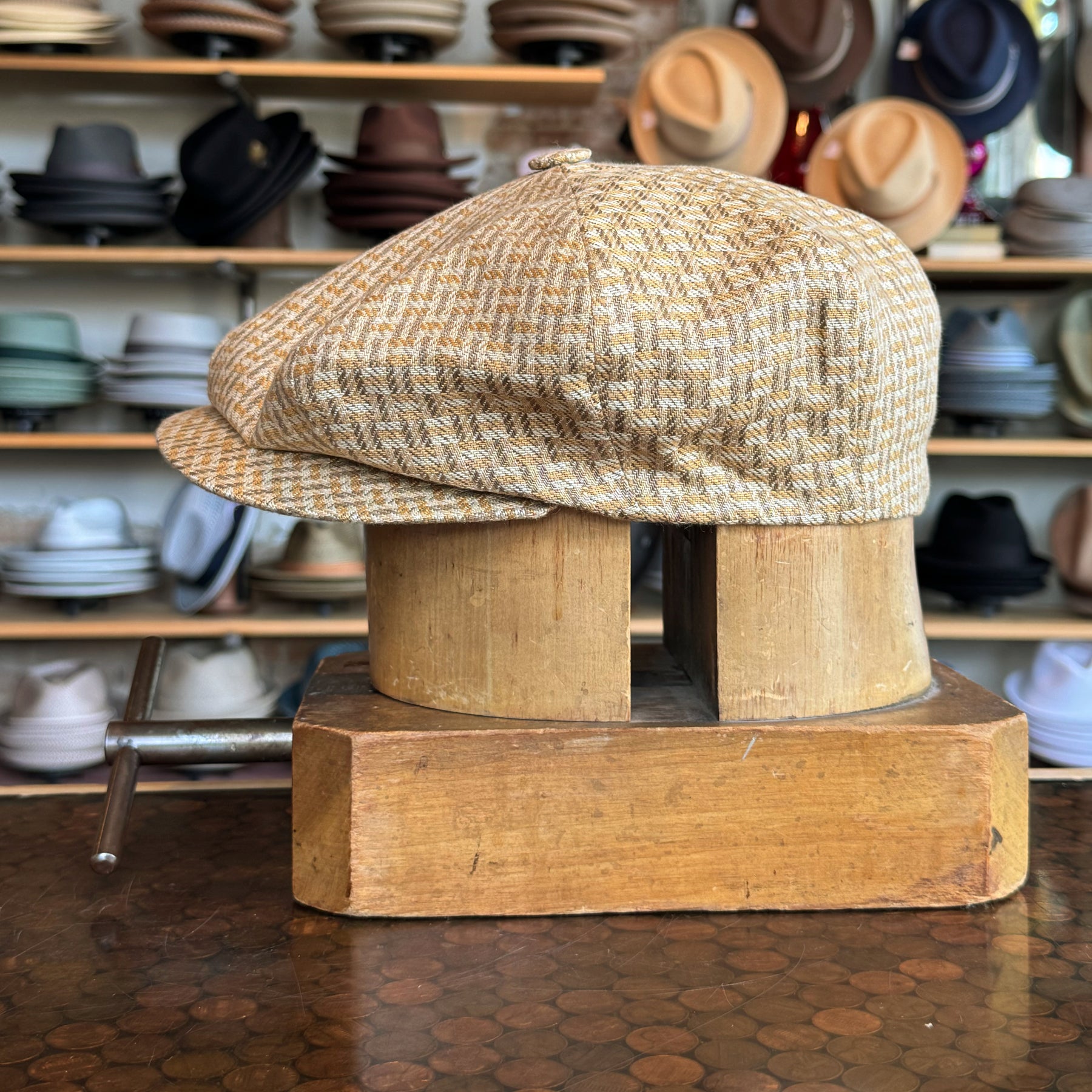 Tetris Jacquard Newsboy cap in a warm golden beige pattern, displayed on a wooden hat block with various hats in the background, showcasing a classic newsboy style.