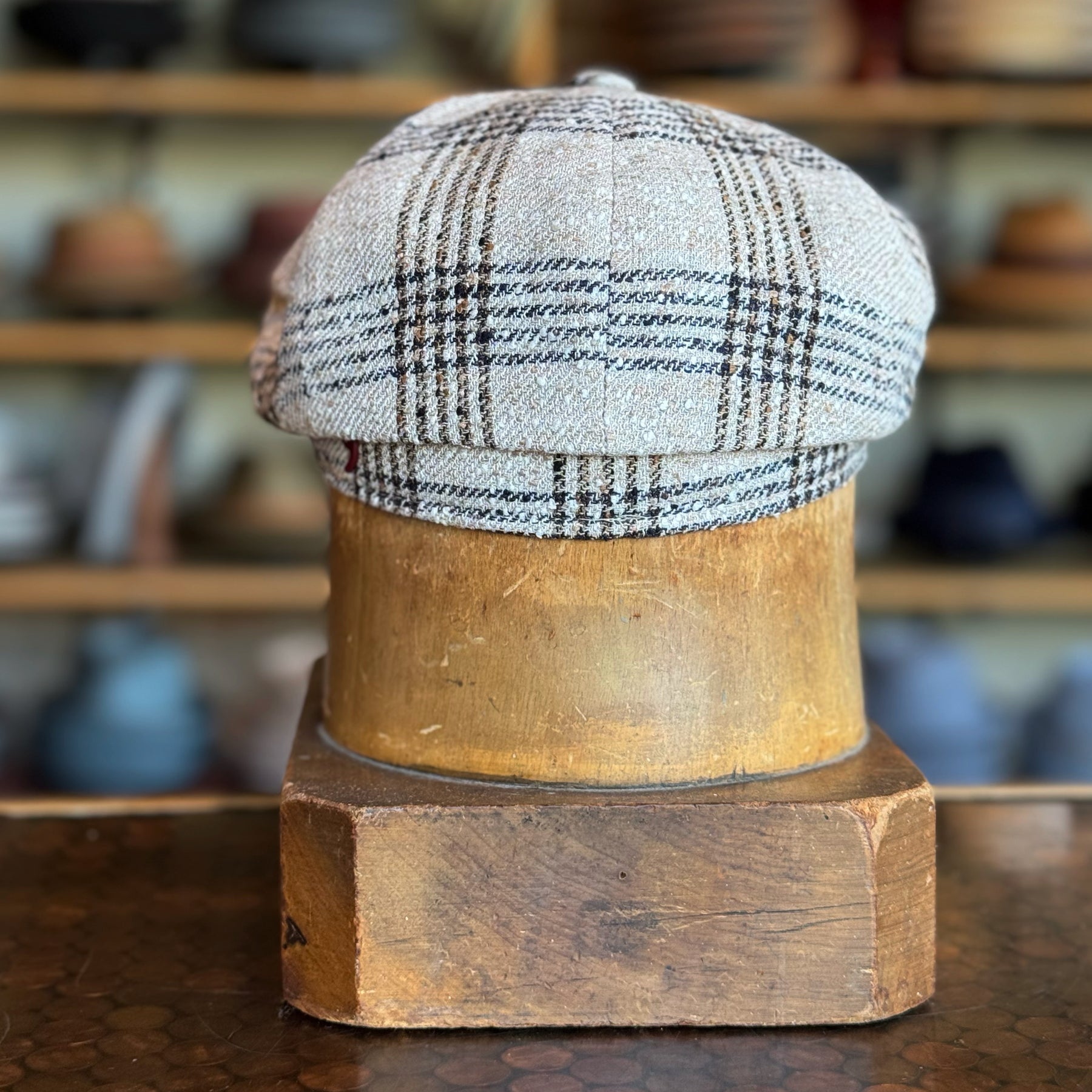 Textured Linen Hatteras flat cap with a plaid pattern in neutral tones displayed on a wooden hat stand in a hat shop setting.