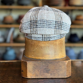 Textured Linen Hatteras flat cap with a plaid pattern in neutral tones displayed on a wooden hat stand in a hat shop setting.